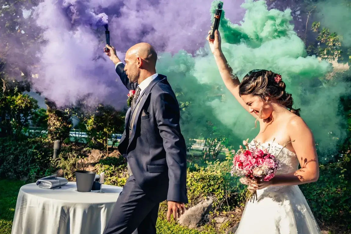 Novios vestidos de gala con bengalas de humo de colores verde y púrpura en la entrada de la ceremonia.