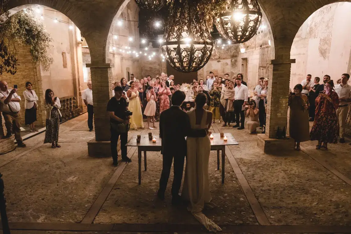 Novios de espaldas, rodeados de invitados en patio iluminado con luces cálidas y ambiente romántico.