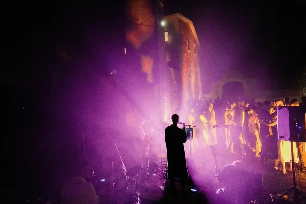 Persona cantando o hablando al micrófono frente a luces y humo rosa, público al fondo.